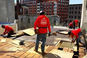 Roofer in a red shirt standing on a flat roof where plywood decking is being replaced, city behind