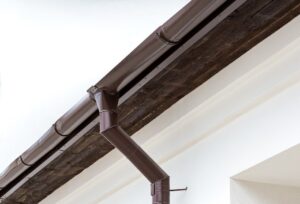 Brown gutter and downspout elbow running along a white soffit against a bright sky