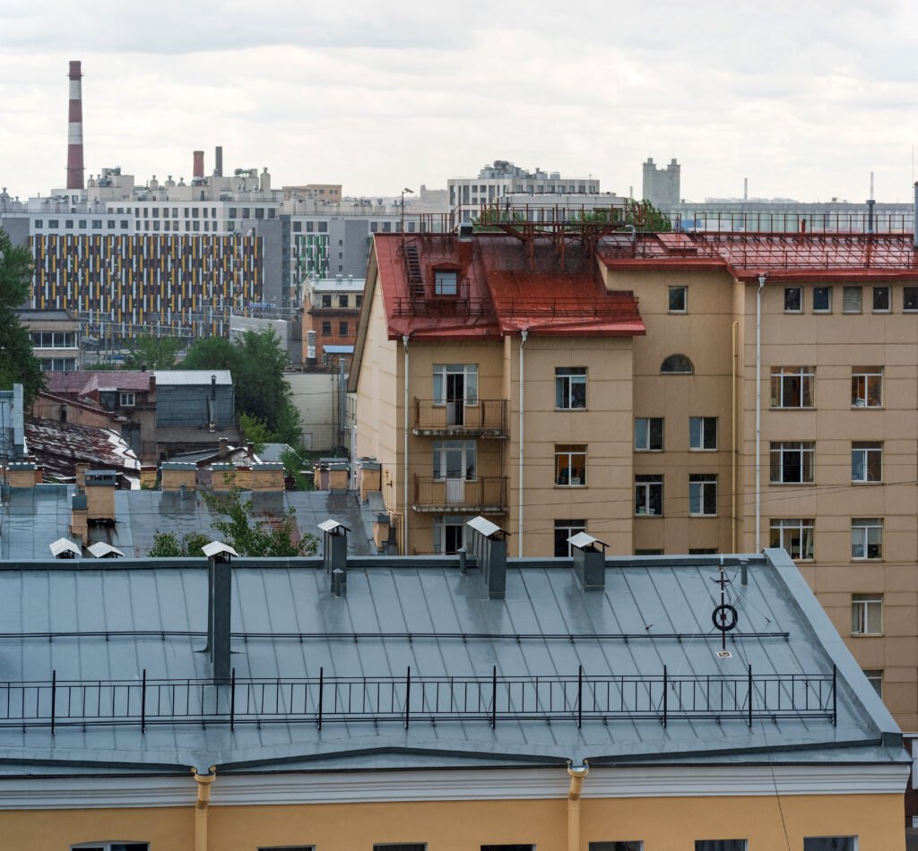 View of city buildings with a red-roofed yellow apartment building in the foreground, a blue-gray roof below, and modern high-rises and a smokestack in the distant background under a cloudy sky.