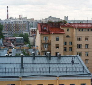 View of city buildings with a red-roofed yellow apartment building in the foreground, a blue-gray roof below, and modern high-rises and a smokestack in the distant background under a cloudy sky.