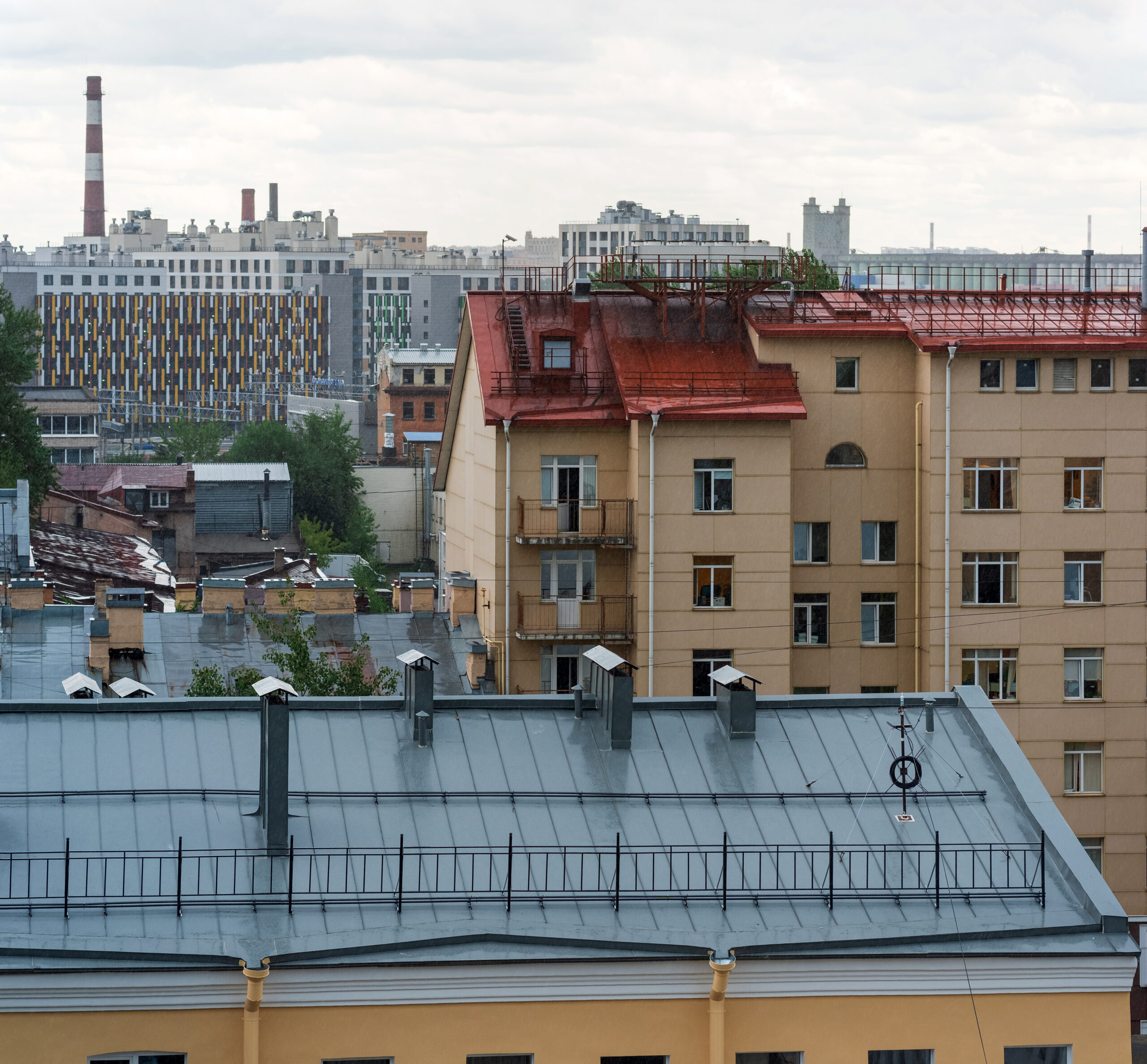 View of city buildings with a red-roofed yellow apartment building in the foreground, a blue-gray roof below, and modern high-rises and a smokestack in the distant background under a cloudy sky.