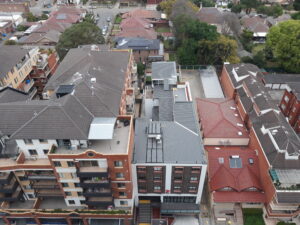 Aerial view of a residential neighborhood with multi-story apartment buildings featuring balconies, red and gray rooftops, and some greenery among the structures. Streets and parked cars can be seen between the buildings.