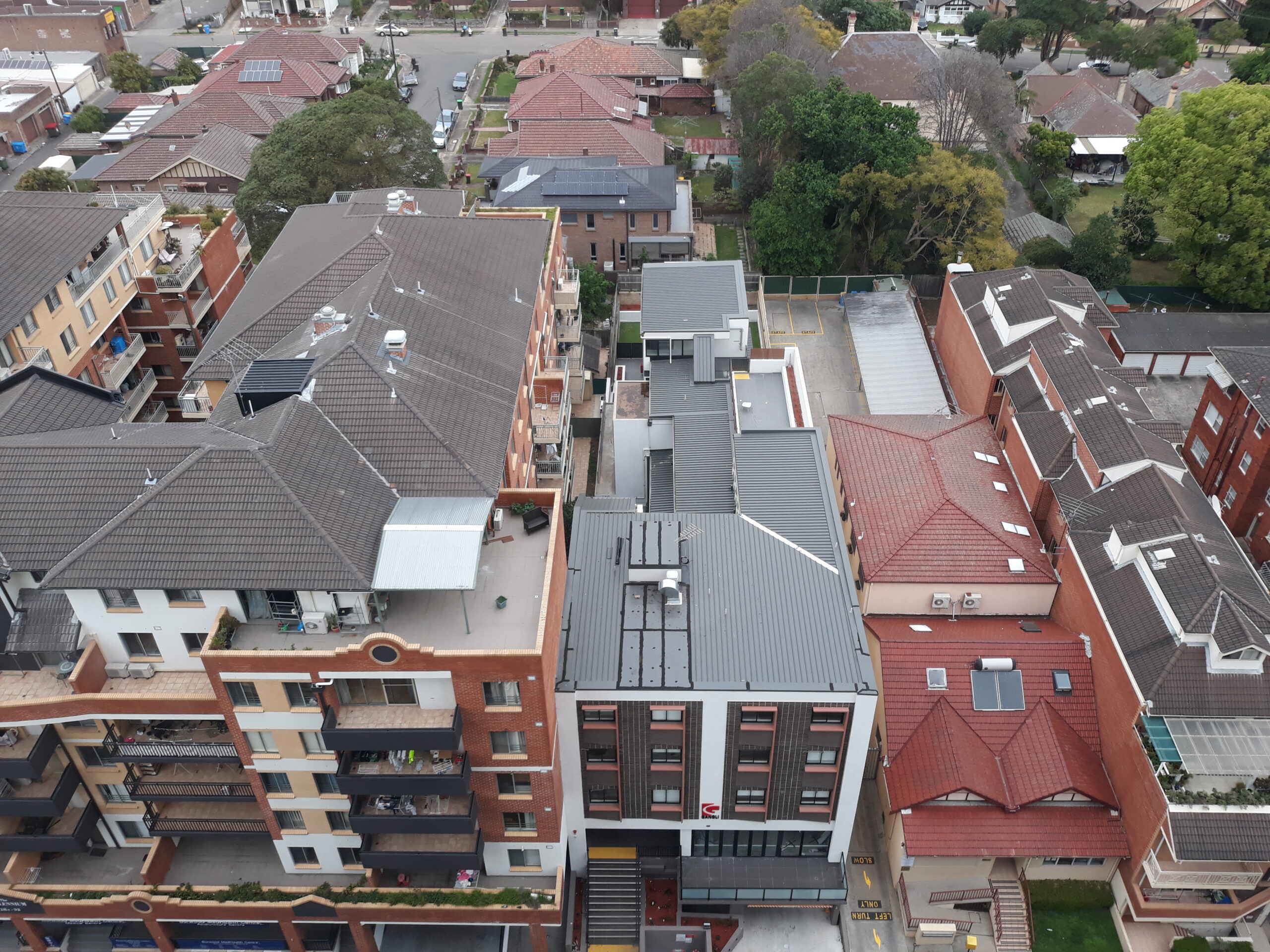 Aerial view of a residential neighborhood with multi-story apartment buildings featuring balconies, red and gray rooftops, and some greenery among the structures. Streets and parked cars can be seen between the buildings.