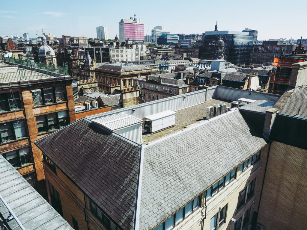 View of city rooftops and historic buildings under a clear blue sky, with modern high-rise structures in the background. The scene combines old and new architectural styles.