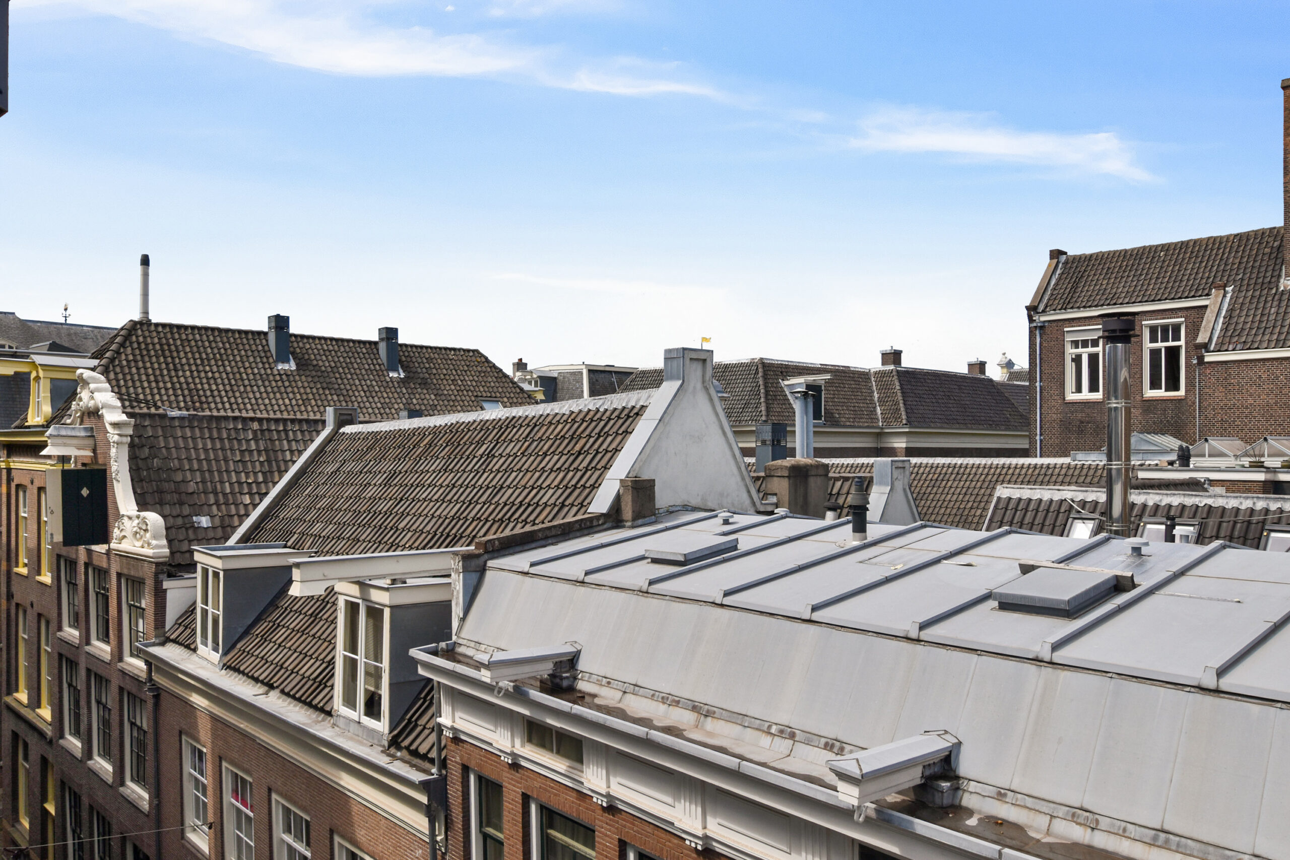 View of traditional European rooftops with red and gray tiles, chimneys, and dormer windows under a bright blue sky with light clouds. The buildings are closely packed, showing a classic urban cityscape.