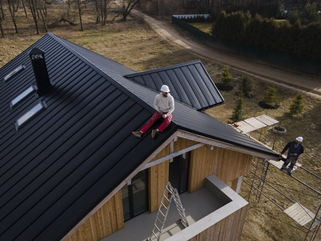 Two construction workers wearing hard hats work on the roof of a modern wooden house. One worker sits near the roof edge, while the other stands on scaffolding, with ladders and construction materials nearby.