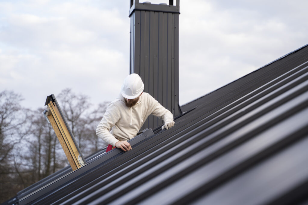 A construction worker wearing a white hard hat and light-colored hoodie is installing or inspecting metal panels on a sloped black metal roof near a chimney on a cloudy day. Trees are visible in the background.