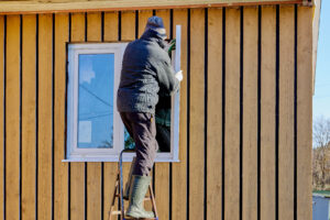 A person in winter clothing stands on a step ladder and installs or repairs a window on the exterior wall of a wooden house.