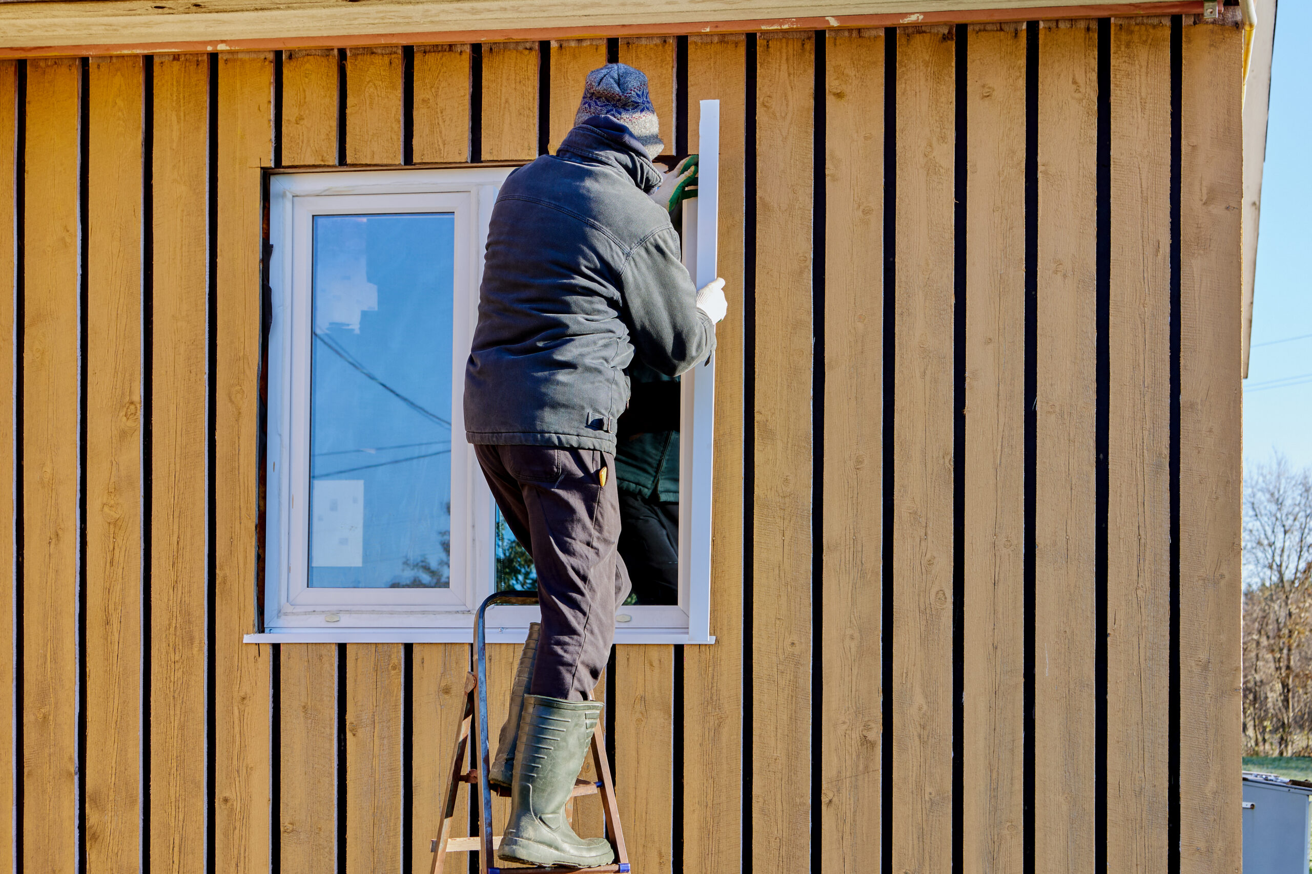 A person in winter clothing stands on a step ladder and installs or repairs a window on the exterior wall of a wooden house.