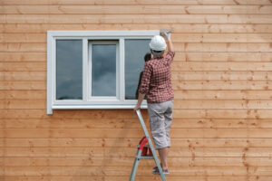 A person wearing a hard hat and plaid shirt stands on a ladder, painting or working on the exterior wall of a wooden house near a white-framed window.